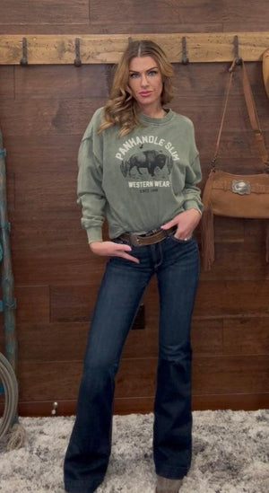 Woman wearing a green sweatshirt with a graphic design and dark jeans, standing against a wooden wall.