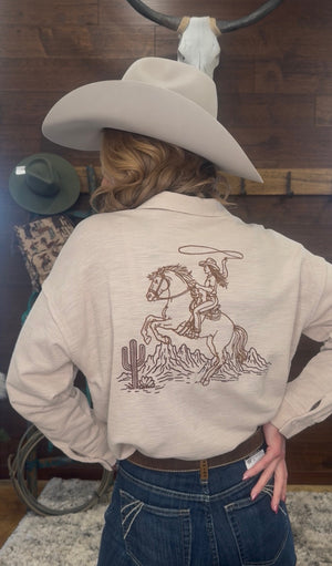 Person wearing a beige jacket with a graphic design of a cowgirl on a horse, standing in a room with wooden walls.
