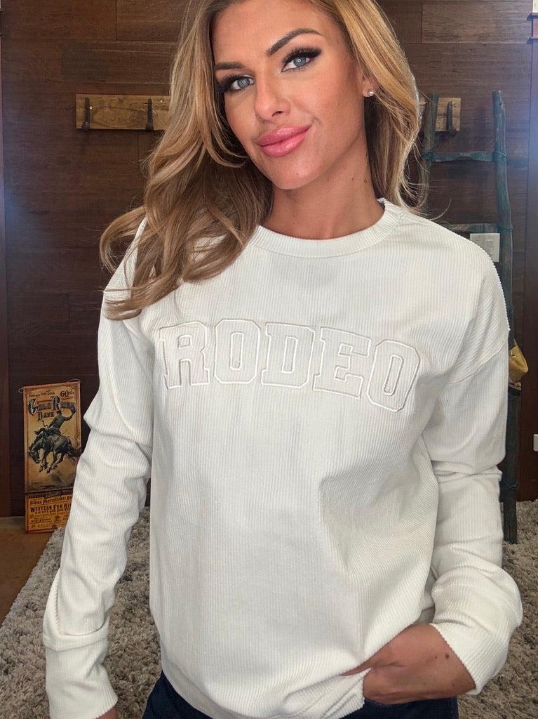 A woman wearing a white long-sleeve crewneck sweatshirt with 'RODEO' printed in tonal arched letters across the chest, paired with dark jeans, standing in front of a rustic wooden wall.