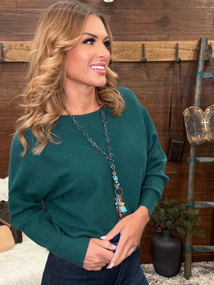 A woman with blonde wavy hair wearing a dark green long-sleeve knit sweater and a long silver chain necklace with colorful beads, standing in front of a rustic wooden wall.