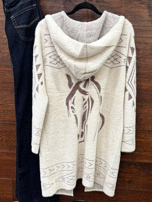 A cream-colored fuzzy hooded cardigan sweater with brown horse and geometric patterns, hanging on a black hanger next to a pair of dark blue jeans against a wooden background.