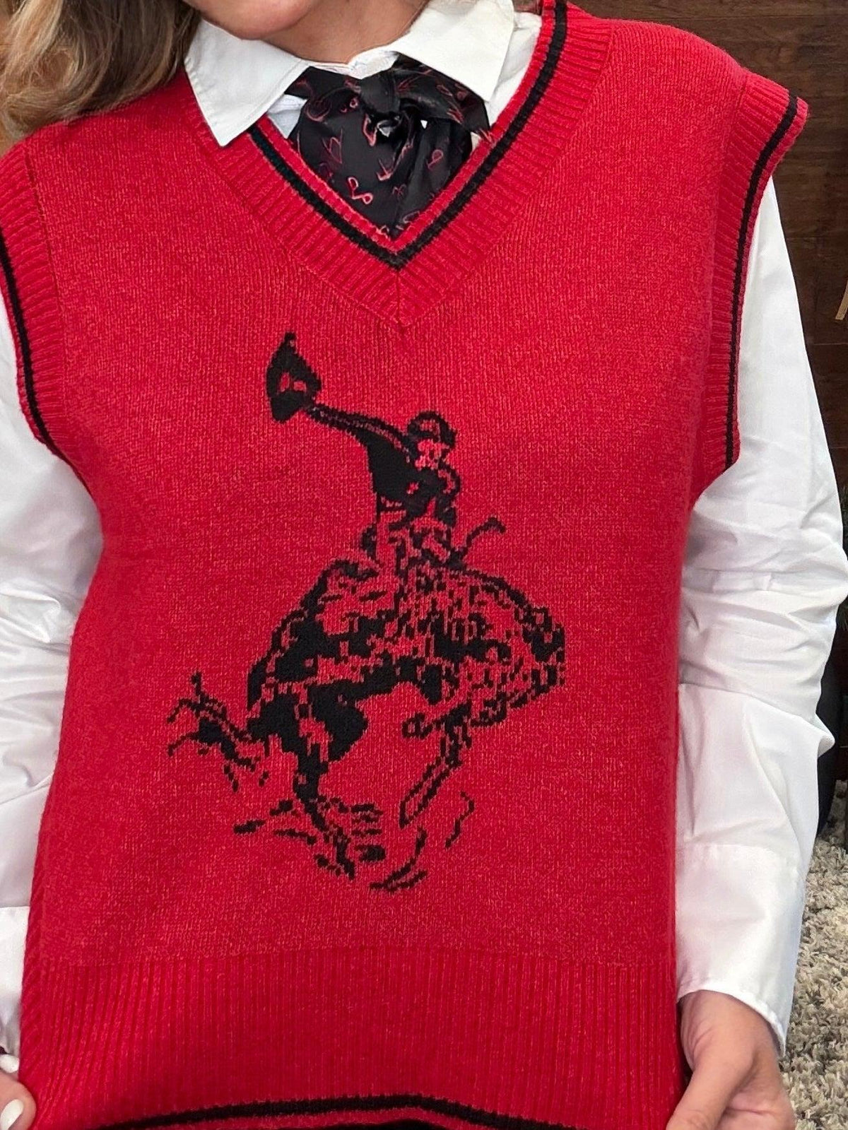A red knit sweater vest with black trim and a black cowboy riding a bucking horse graphic, worn over a white collared shirt and black bow tie.