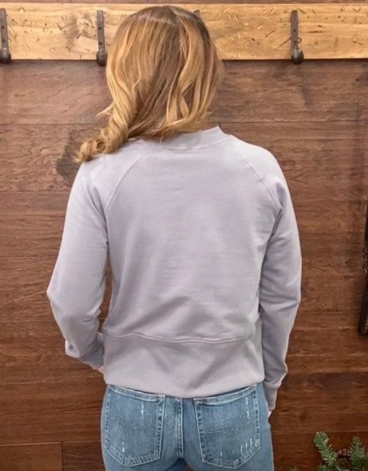 A woman wearing a light gray long-sleeve crewneck sweatshirt with 'ARIAT' printed across the chest.