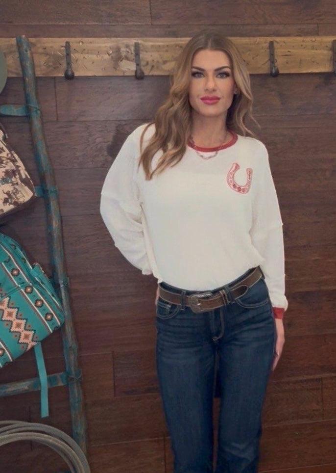Woman wearing a white sweatshirt with a red horseshoe design and blue jeans, standing against a wooden wall.