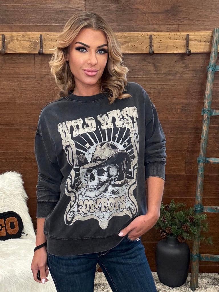 A woman wearing a faded black graphic sweatshirt with a western skull design and the words 'Wild West Cowboys', paired with dark blue jeans, standing in front of a rustic wooden wall.