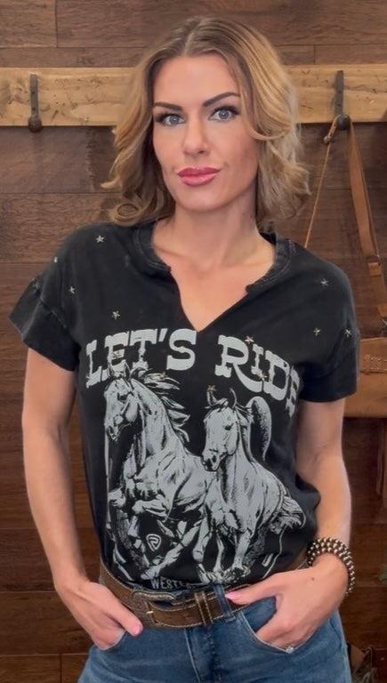 Woman wearing a black t-shirt with horse graphic and 'Let's Ride' text in a rustic setting.