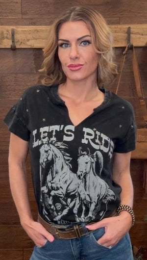 Woman wearing a black t-shirt with horse graphic and 'Let's Ride' text in a rustic setting.