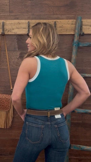 Woman wearing a teal tank top and blue jeans standing in front of a wooden wall.