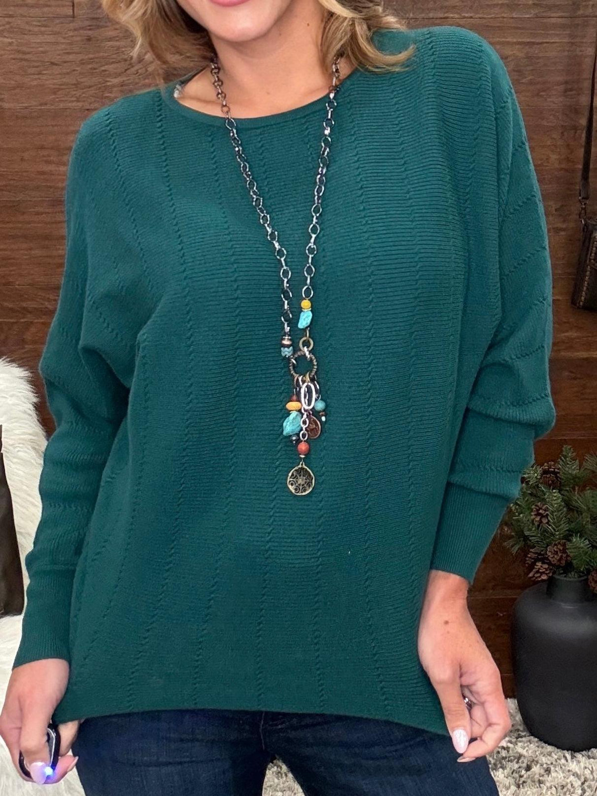A woman wearing a dark teal green long-sleeve knit sweater with a textured cable pattern, paired with a long silver chain necklace with colorful beads and charms, and dark blue jeans.