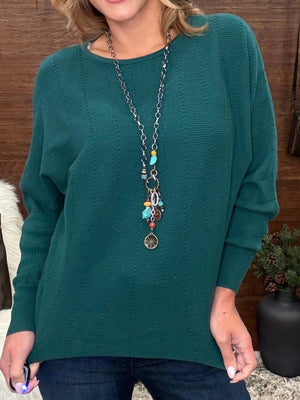 A woman wearing a dark teal green long-sleeve knit sweater with a textured cable pattern, paired with a long silver chain necklace with colorful beads and charms, and dark blue jeans.