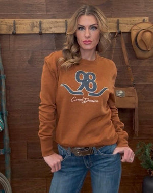 Woman wearing a brown sweatshirt with a logo and text, standing against a wooden wall.