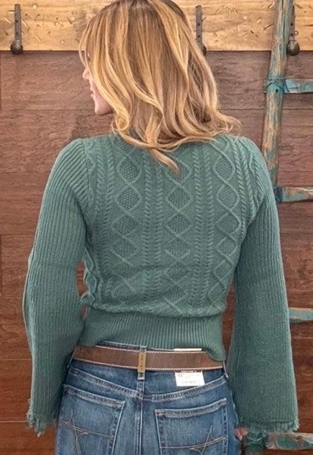A woman viewed from behind wearing a sage green cable-knit sweater and blue jeans, standing in front of a rustic wooden wall.