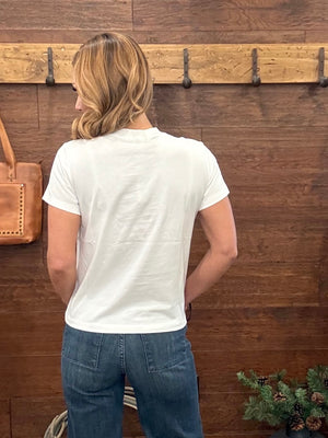 A woman viewed from behind wearing a plain white short-sleeve t-shirt and blue jeans, standing in front of a rustic dark wood wall with a wooden coat rack.