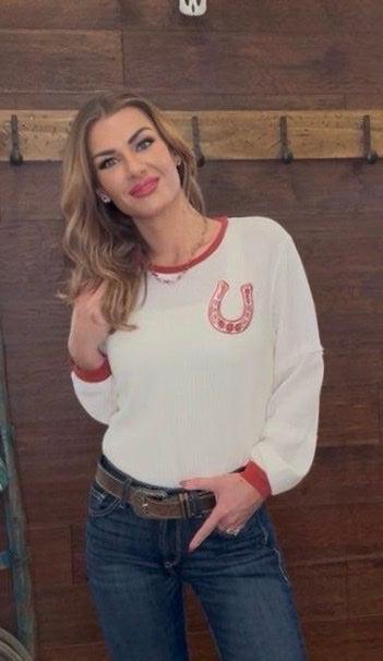 Woman wearing a white sweater with a red horseshoe design in front of a wooden wall.