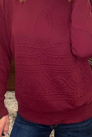 A close-up of a person wearing a burgundy long-sleeve sweater with raised decorative knit patterns across the chest and waist, paired with blue jeans.