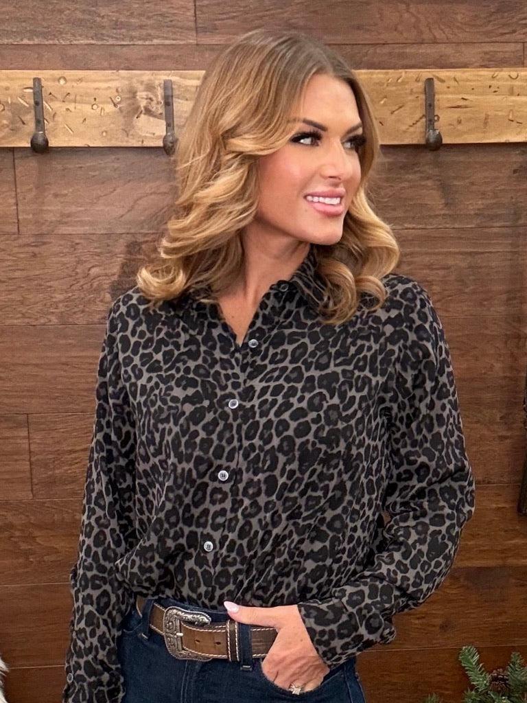 A woman wearing a black and gray leopard print button-down shirt, styled with dark blue jeans and a brown leather belt with a silver buckle, standing in front of a rustic wooden wall.
