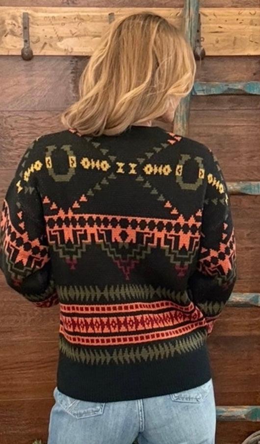 A woman viewed from behind wearing a black knit sweater with a colorful geometric pattern in orange, red, and olive green, paired with light blue jeans, standing against a rustic wooden wall.
