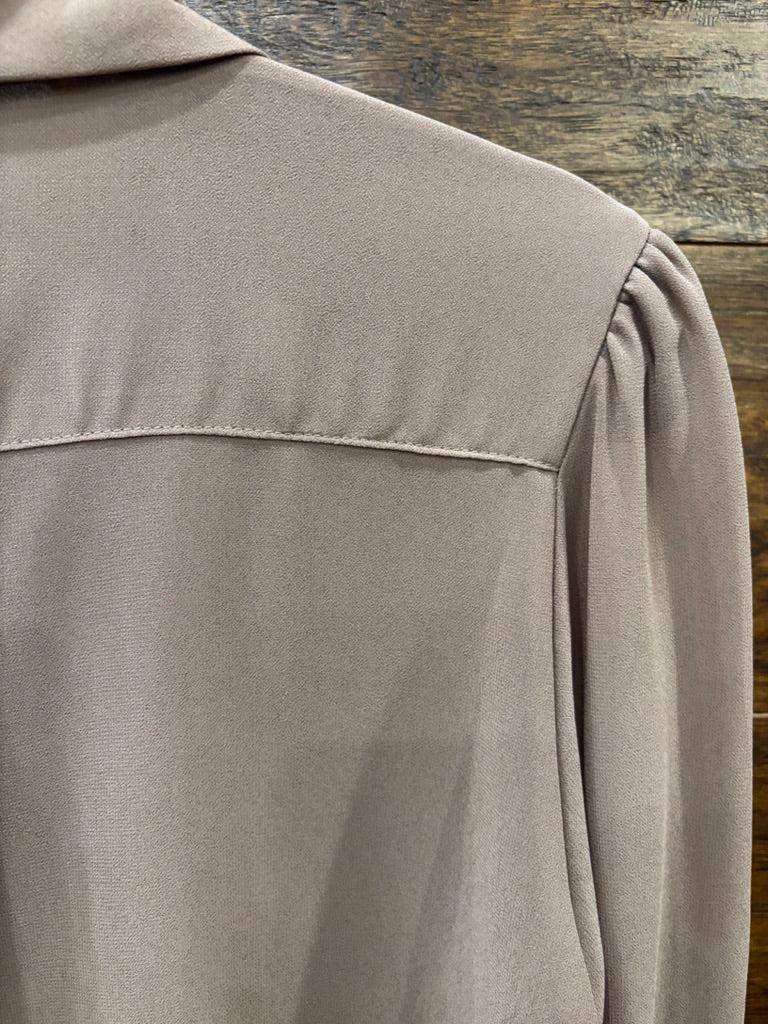 A taupe long-sleeve button-down blouse hanging on a black hanger against a rustic wooden wall, styled with dark blue jeans and a cream crossbody bag on hooks.
