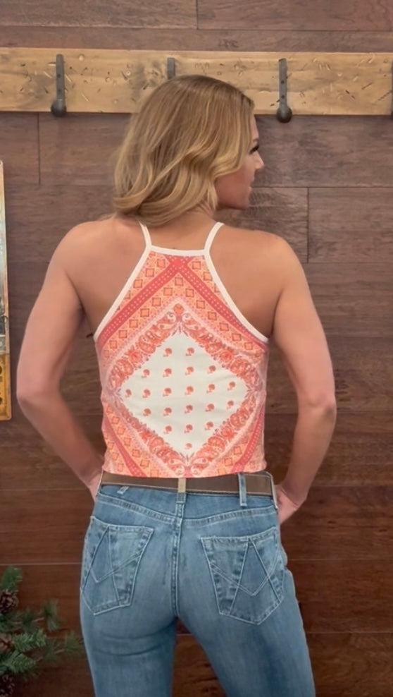 Woman wearing a patterned tank top and blue jeans against a wooden wall.