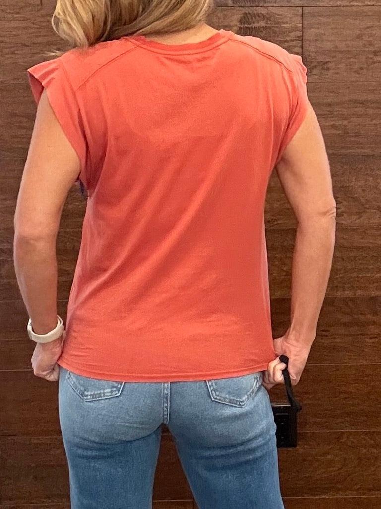 A woman viewed from behind wearing a coral orange short-sleeve t-shirt and light blue jeans, standing against a dark wood-paneled wall.
