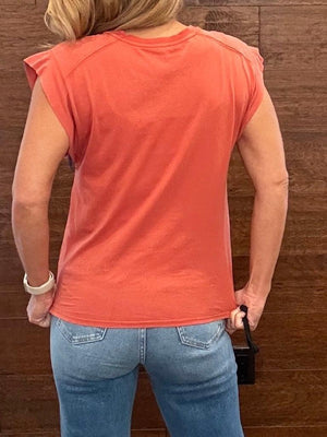 A woman viewed from behind wearing a coral orange short-sleeve t-shirt and light blue jeans, standing against a dark wood-paneled wall.