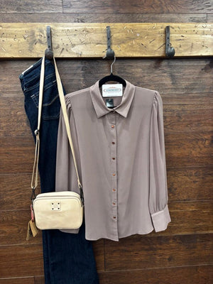 A taupe long-sleeve button-down blouse hanging on a black hanger against a rustic wooden wall, styled with dark blue jeans and a cream crossbody bag on hooks.