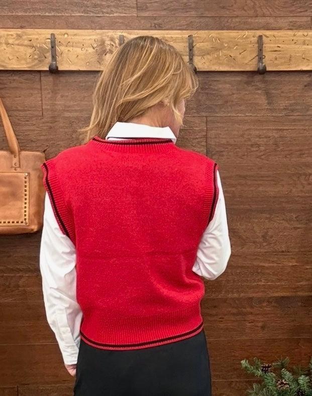A woman viewed from behind wearing a red ribbed knit sweater vest over a white collared shirt and a black skirt, standing against a rustic wooden wall with hooks.