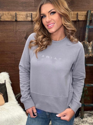 A woman wearing a light gray long-sleeve crewneck sweatshirt with 'ARIAT' printed across the chest.