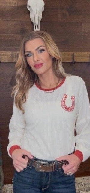 Woman wearing a white sweater with a red horse shoe design, standing against a wooden wall with a skull.