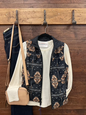 A sleeveless black quilted vest with a tan and brown Southwestern geometric pattern, layered over a cream waffle-knit long-sleeve shirt.