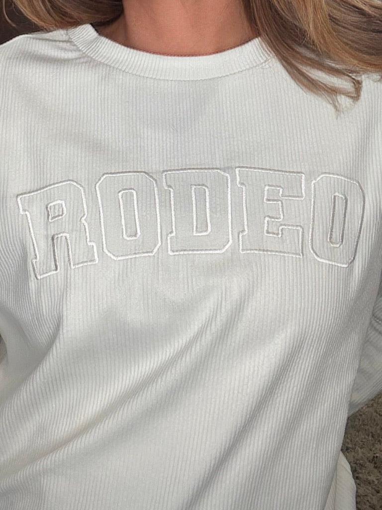 A close-up of a person wearing a ribbed white long-sleeve t-shirt with 'RODEO' embroidered in arched varsity block letters across the chest.