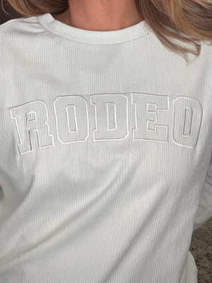 A close-up of a person wearing a ribbed white long-sleeve t-shirt with 'RODEO' embroidered in arched varsity block letters across the chest.
