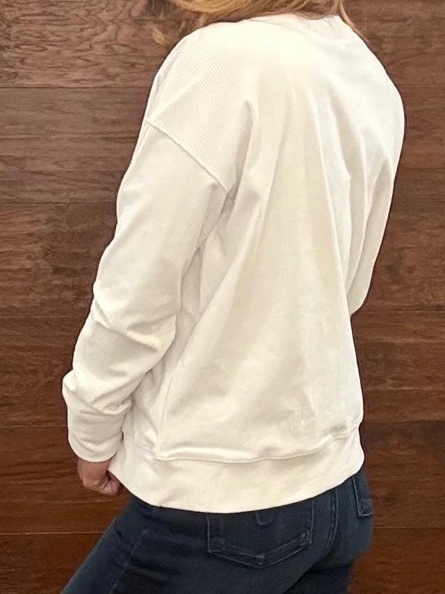 A woman wearing a white long-sleeve crewneck sweatshirt with 'RODEO' printed in tonal arched letters across the chest, paired with dark jeans, standing in front of a rustic wooden wall.
