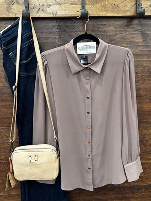 A taupe long-sleeve button-down blouse hanging on a black hanger against a rustic wooden wall, styled with dark blue jeans and a cream crossbody bag on hooks.