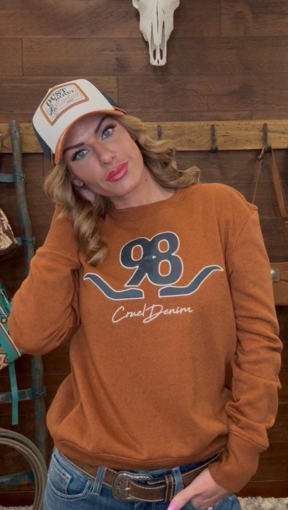 Woman wearing a brown sweatshirt with a logo and text, posing in front of a wooden wall.