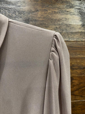 A taupe long-sleeve button-down blouse hanging on a black hanger against a rustic wooden wall, styled with dark blue jeans and a cream crossbody bag on hooks.