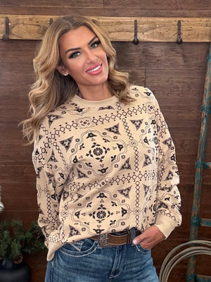 A woman wearing a beige long-sleeve crewneck sweatshirt with a black geometric southwestern-style pattern, paired with blue jeans and a brown belt, standing in front of a rustic wooden wall.