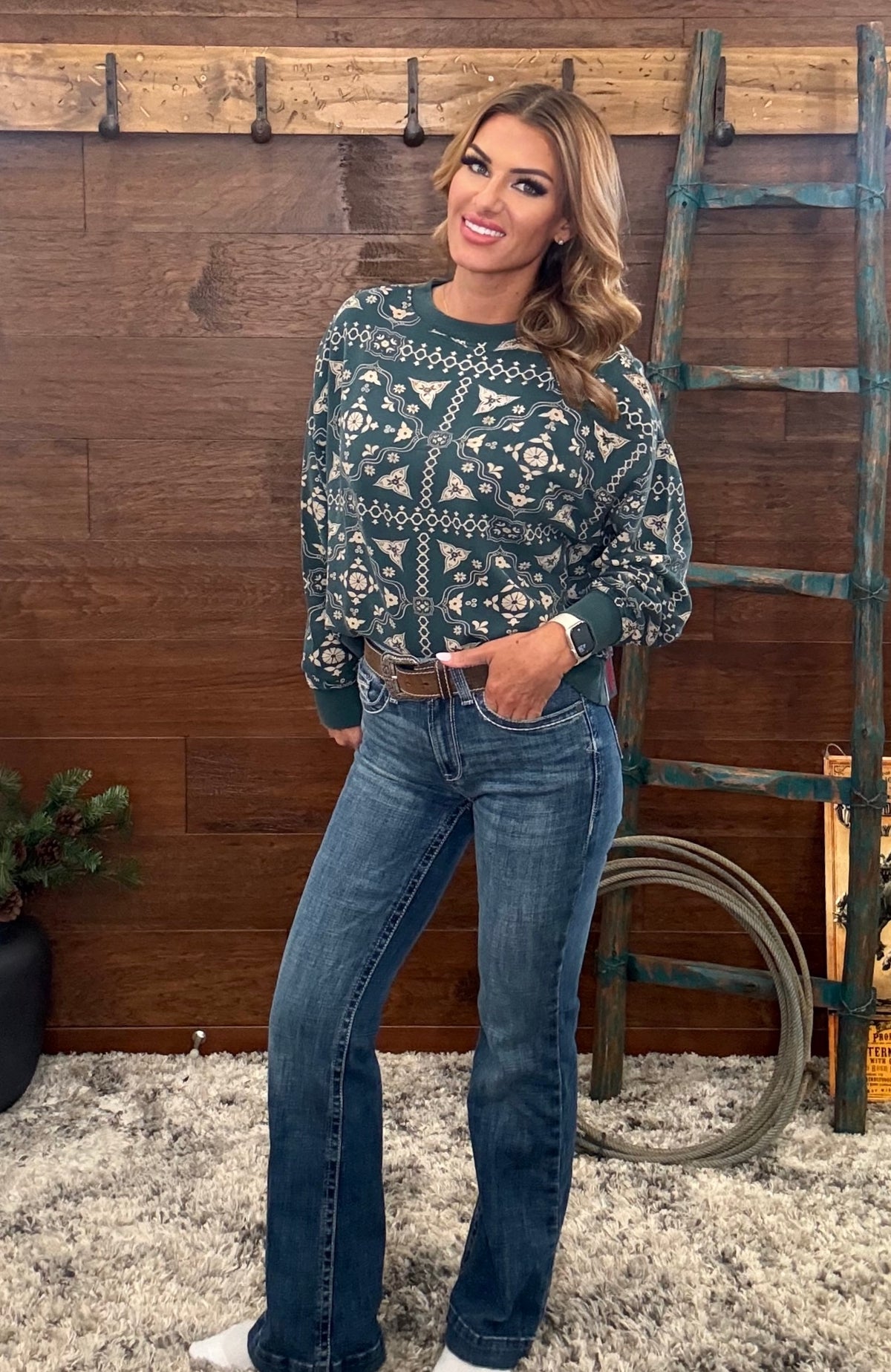 A woman modeling a dark green and white patterned knit sweater with blue jeans, standing in front of a rustic wooden wall with a ladder and coat rack.