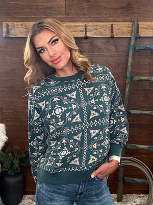 A woman wearing a dark teal green long-sleeve crewneck sweatshirt with a cream-colored geometric and floral pattern, paired with blue jeans, standing in front of a rustic wooden wall.