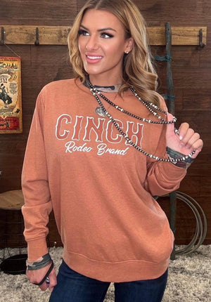 A woman wearing a burnt orange long-sleeve sweatshirt with 'CINCH Rodeo Brand' printed in white across the chest, paired with dark blue jeans and layered silver chain necklaces, posed in front of a rustic wooden wall.