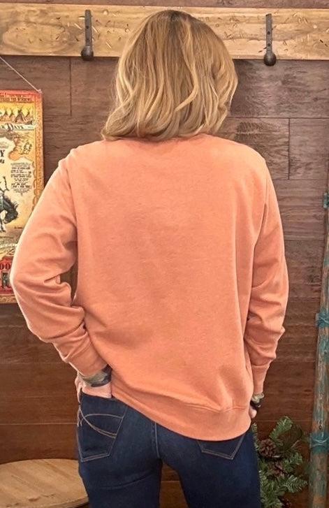 A woman viewed from behind wearing a peach-colored long-sleeve crewneck sweatshirt and dark blue jeans, standing in front of a rustic wooden wall.
