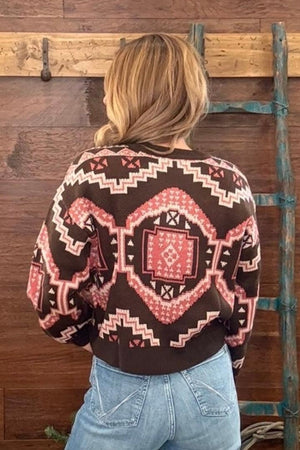 A woman viewed from behind wearing a brown knit sweater with a pink and cream geometric southwestern pattern, paired with light blue jeans, standing in front of a rustic wooden wall.