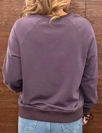 A person viewed from the back wearing a muted mauve-purple crewneck sweatshirt and light blue jeans, standing against a brown wooden wall.