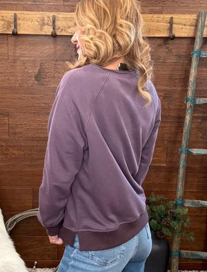 A woman with blonde wavy hair is shown from the back wearing a dusty purple crewneck sweatshirt and light blue jeans, standing in front of a rustic wooden wall.
