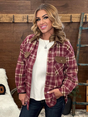 A woman wearing a burgundy and tan plaid button-up shirt jacket over a white ribbed knit top and dark jeans, standing in front of a rustic wood wall with a ladder and faux fur decor.