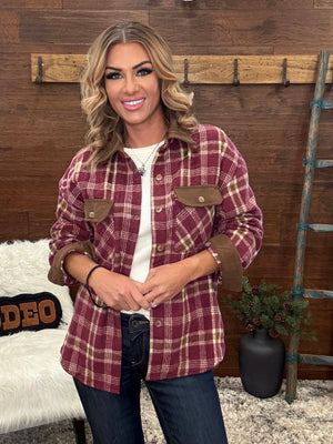 A woman wearing a burgundy and tan plaid flannel button-up shirt over a white top and dark blue jeans, standing in a rustic room with wood paneling, a coat rack, and a ladder.