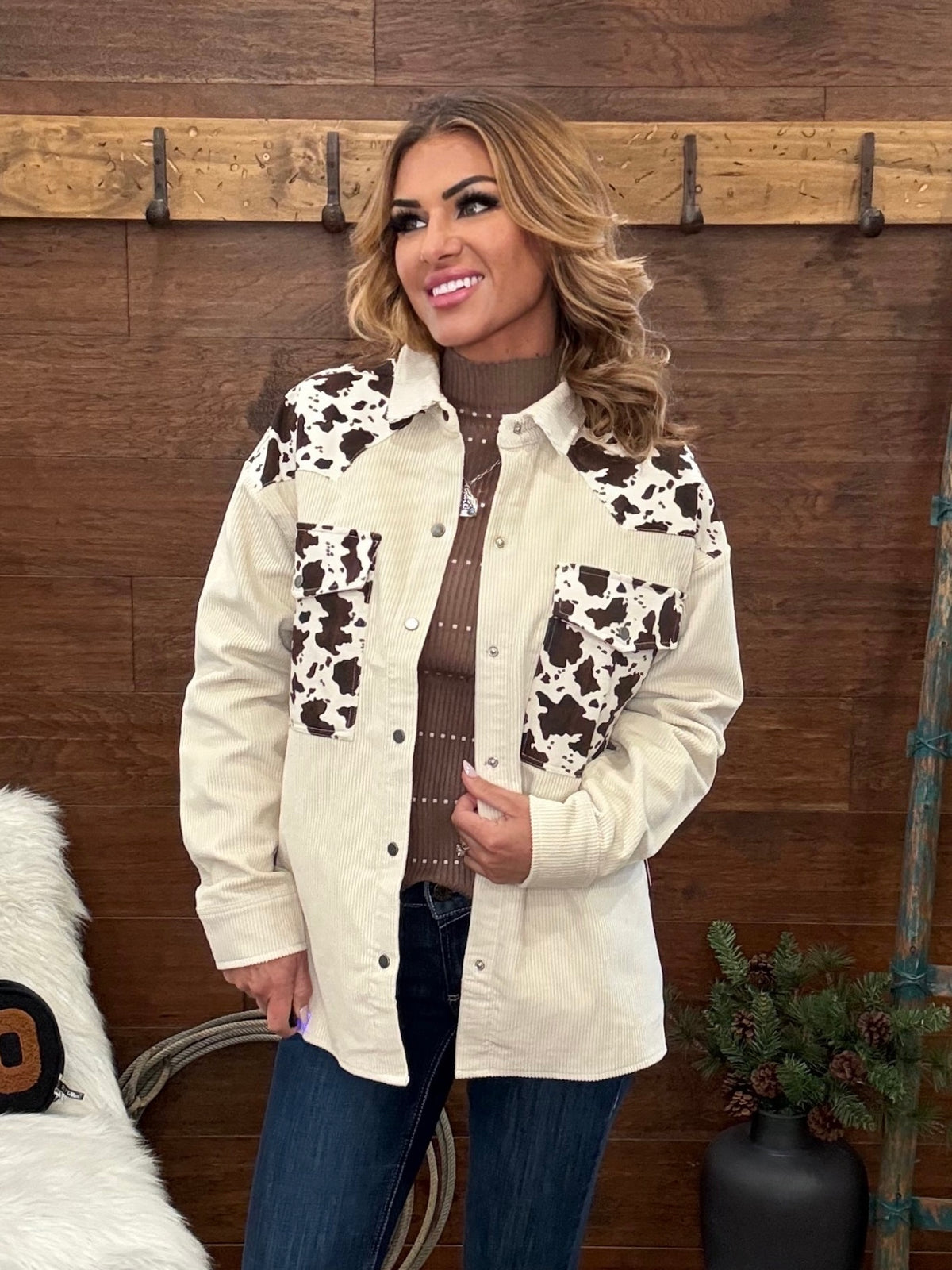 A woman wearing a cream western-style button-up shirt jacket with brown and black cow-print panels on the chest and pockets, layered over a brown ribbed turtleneck and blue jeans, standing in front of a rustic wood wall.