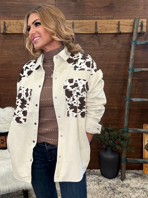 A woman wearing a cream-colored button-up shirt jacket with brown and black cow-print panels on the shoulders, chest, and pockets, layered over a brown ribbed turtleneck sweater and dark blue jeans, standing in front of a rustic wooden wall.