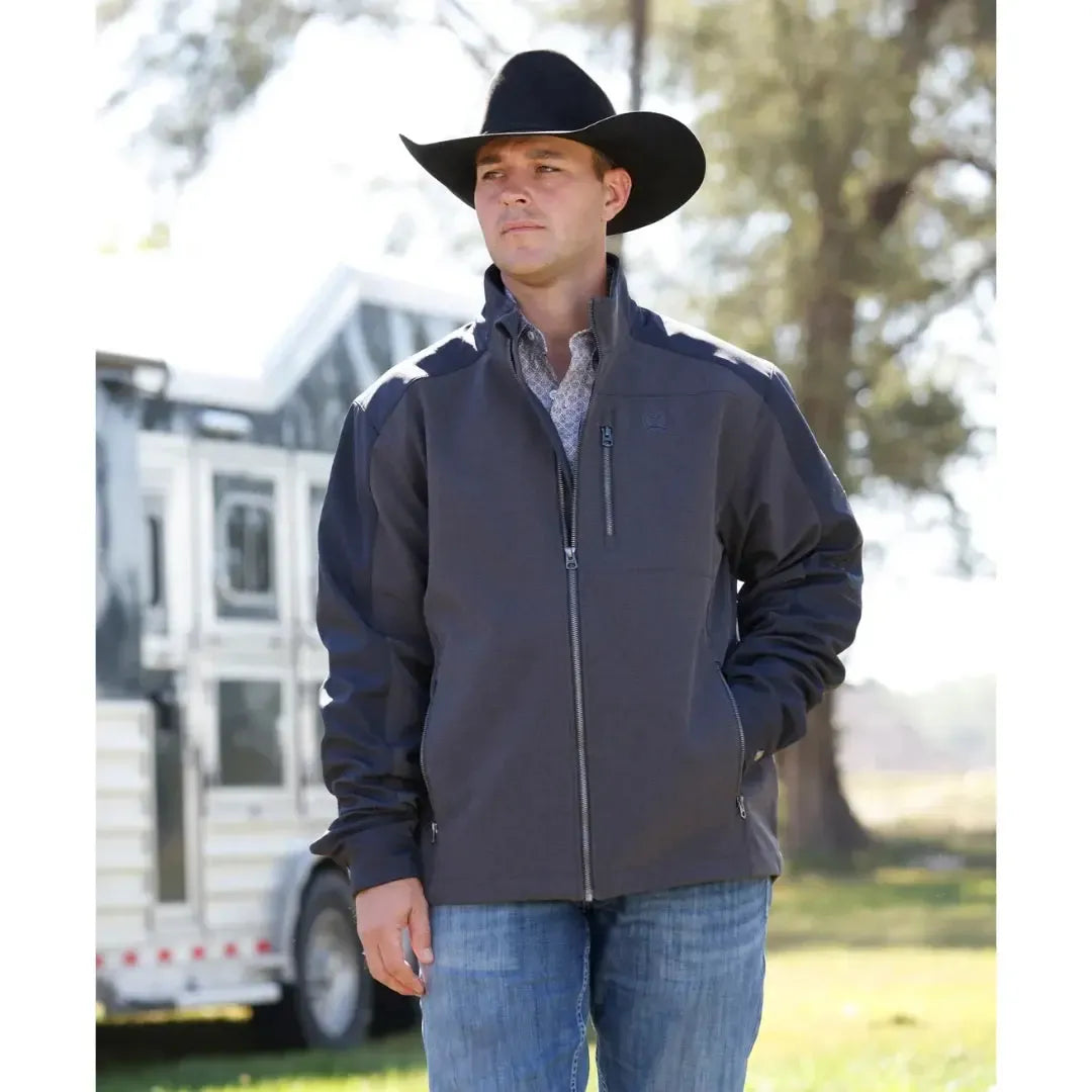 A man wearing a dark gray and black Clinch outdoor jacket with a full-zip front, zippered chest pocket, and vertical Clinch branding on the sleeve, paired with blue jeans against a white background.