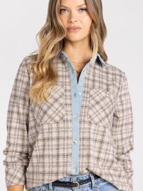 A woman wearing a beige and brown plaid button-up shirt over a light blue collared shirt, paired with blue jeans, against a plain light gray background.